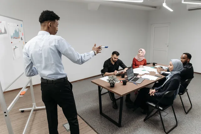 A multicultural business team engaged in a discussion during an office meeting with a whiteboard and laptops. Resolving repeated handoff disagreements