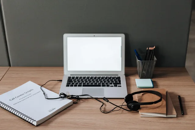 Minimalist workspace with laptop, headset, and call center scripts on a wooden desk. Sales development tools
